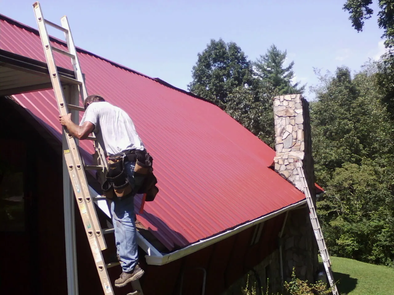 Roofer on a ladder beside a red metal roof and stone chimney — Soffit & Fascia Repair by Roofing Brothers in Highfalls